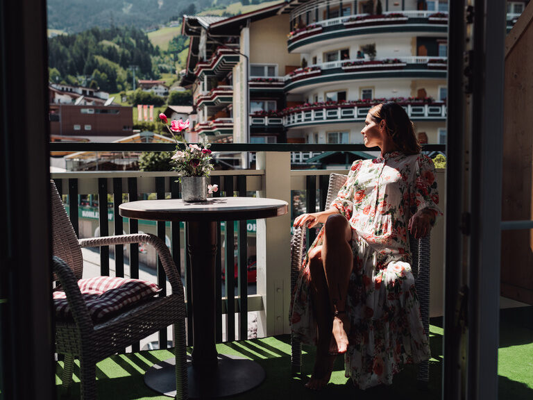 Frau sitzt im Sommerkleid auf dem Zimmerbalkon, genießt die Sonne und blickt auf die Talstation der Spieljochbahn