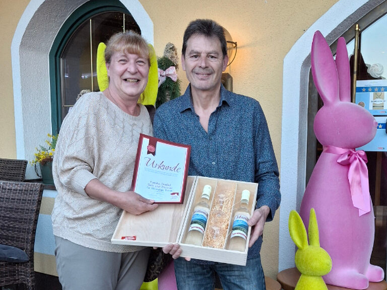 Paar steht vor dem Kohlerhof und posiert mit der Ehrenurkunde und Geschenk von den Gastgebern; dahinter ist Osterdeko zu sehen