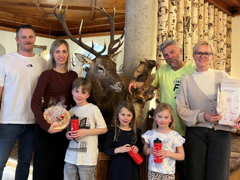 zwei Familien bestehend aus ingesamt vier Erwachsenen und drei Kindern stehend lächelnd vor dem ausgestopften Hirsch und halten die Geschenke vom Hotel in den Händen