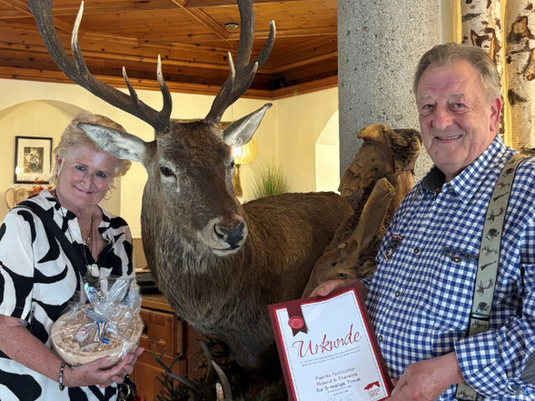 Paar posiert mit Urkunde und Geschenk anlässlich Gästeehrung in der Hotellobby des Kohlerhofs