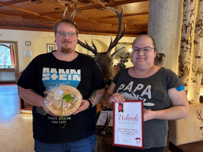 Paar steht in der Lobby und posiert mit Ehrenurkunde und Geschenk vom Hotel Kohlerhof