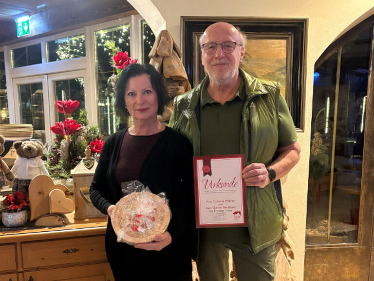 Paar hält Stammgast-Urkunde und Geschenk vom Hotel in den Händen und lächelt in die Kamera, dahinter ist weihnachtliche Deko zu sehen