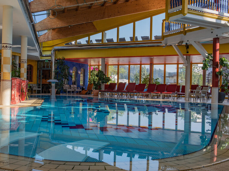 Erlebnishallenbad und Liegestühle, dahinter Glasfront mit Blick auf die Berge