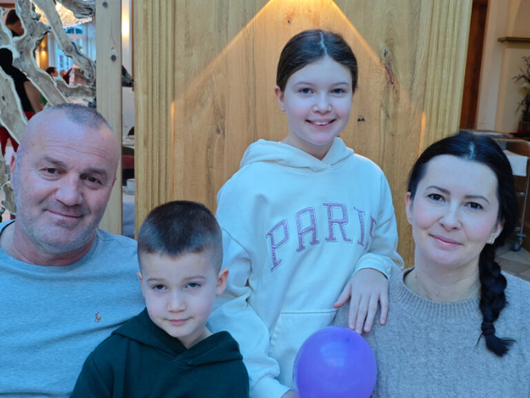 Familie sitzt am Geburtstagstisch und lächelt in die Kamera, ein violetter Luftballon ist ebenfalls zu sehen