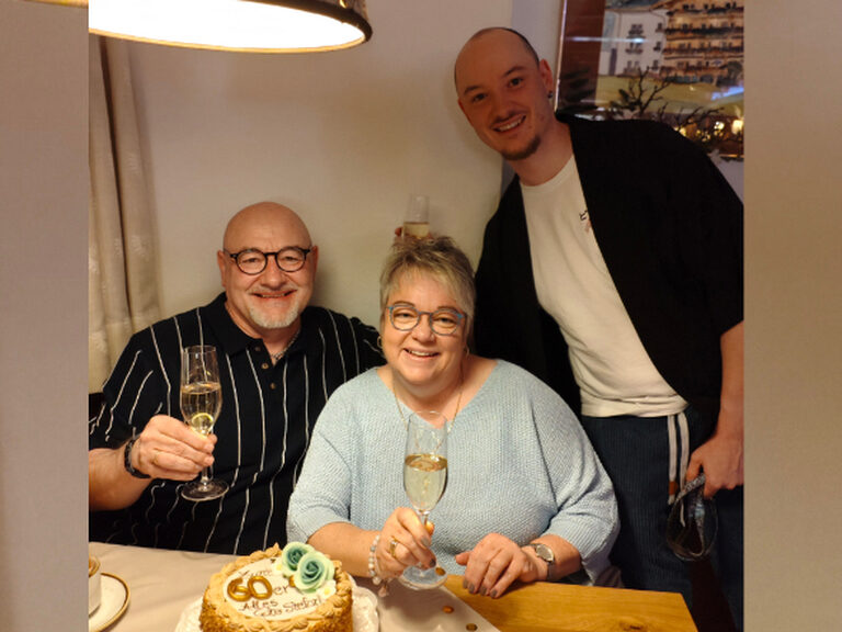 Drei Personen sitzen am festlich gedeckten Geburtstagstisch und stoßen auf einen sechzigsten Geburtstag an