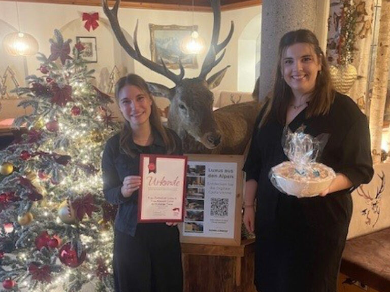 zwei junge Frauen stehen festlich gekleidet vor dem Weihnachtsbaum und halten ihr Geschenk und ihre Gästeehrungsurkunde in den Händen
