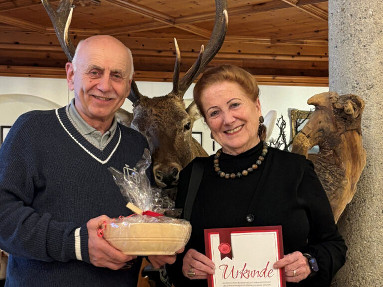 Paar posiert lächelnd mit Gästeurkunde und Geschenk vom Hotel Kohlerhof