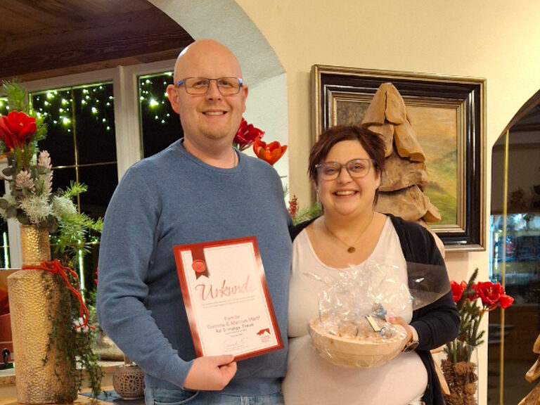 Paar lächelt anlässlich Gästeehrung mit Urkunde und Geschenk vom Hotel in die Kamera
