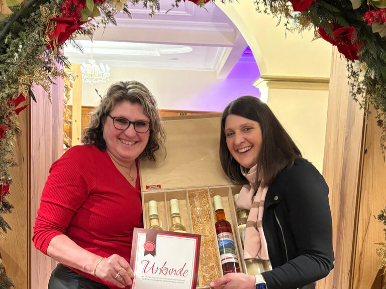 zwei Frauen posieren unter einem Blumenbogen mit Urkunde anlässlich Gästeehrung