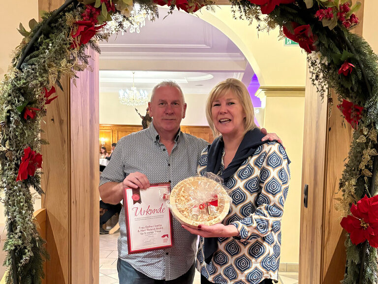 Paar posiert mit Urkunde anlässlich Gästeehrung unter dekoriertem Bogen