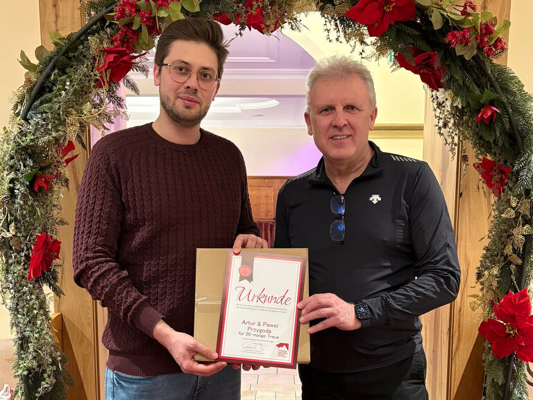 zwei Männer posieren mit Urkunde anlässlich Gästeehrung unter dekoriertem Bogen