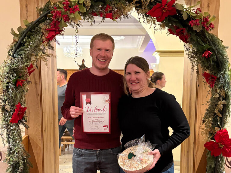 Paar posiert mit Urkunde anlässlich Gästeehrung unter dekoriertem Bogen