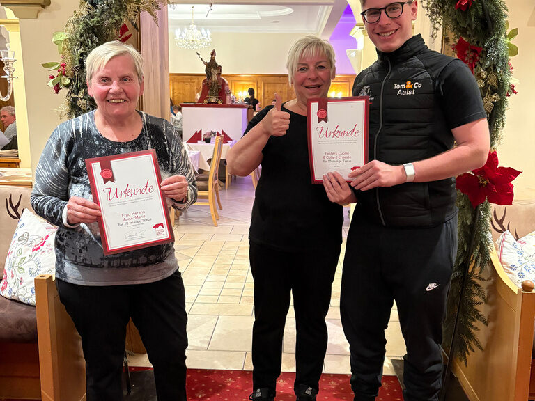 drei Personen posieren mit Urkunde anlässlich Gästeehrung unter dekoriertem Bogen