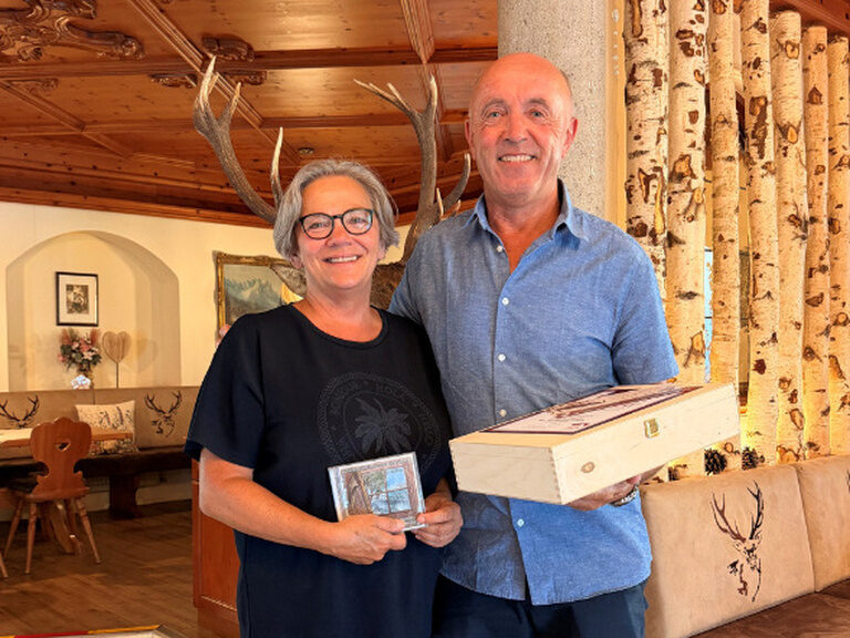 Paar posiert lächelnd in der Kohlerhof-Lobby mit dem Geschenk und der Urkunde des Hotels