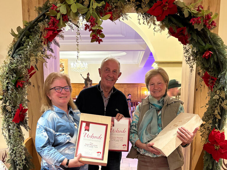 drei Personen posieren mit Urkunde anlässlich Gästeehrung unter dekoriertem Bogen