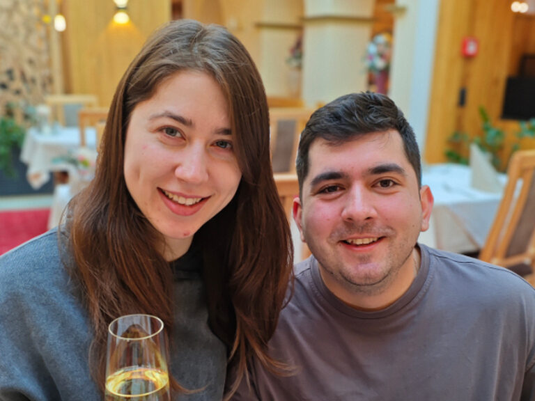 Paar sitzt auf einer Bank und lächelt mit einem Glast Sekt in der Hand anlässlich des Geburtstags der Frau in die Kamera