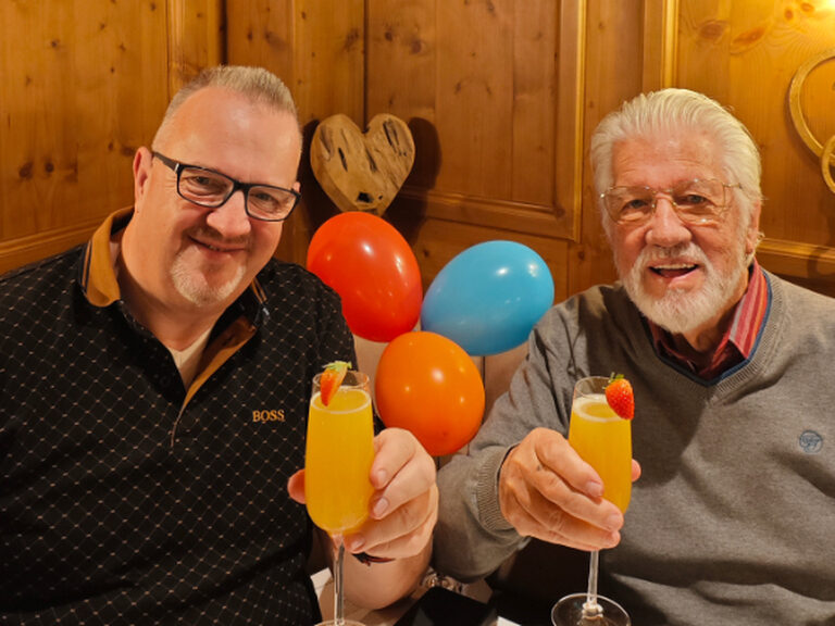 zwei Männer stoßen auf einen Geburtstag an, im Hintergrund sind bunte Luftballone zu sehen