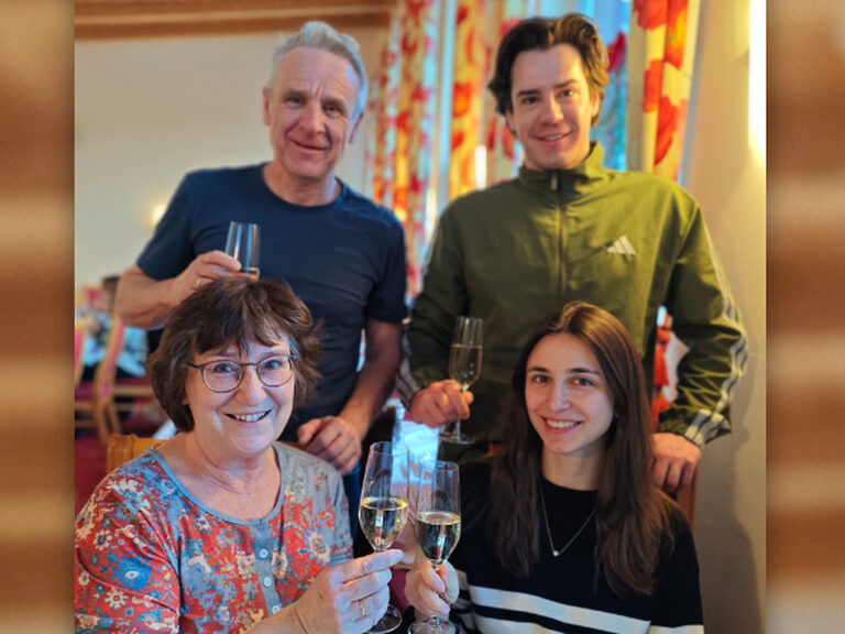 zwei Männer stehen und zwei Frauen sitzen am Tisch und stoßen anlässlich eines Geburtstags an