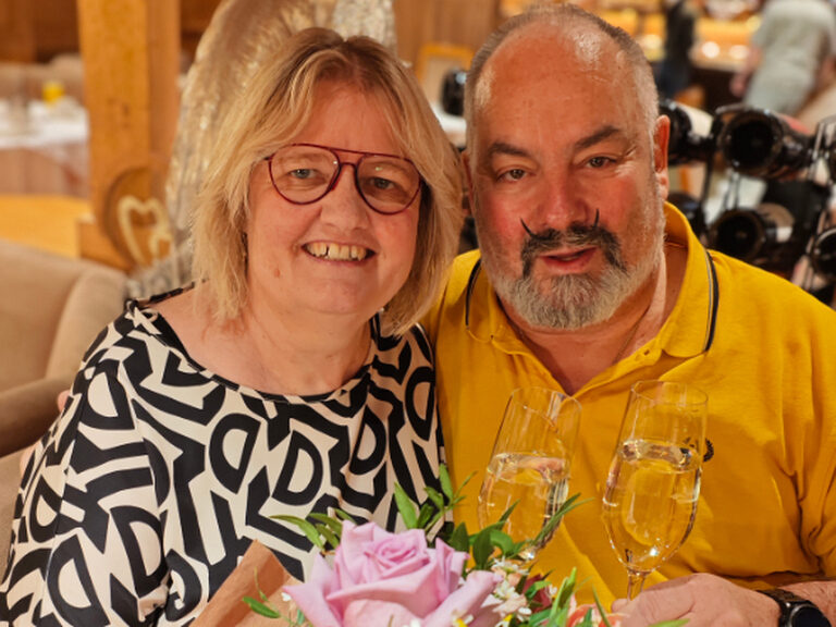 Paar sitzt und lächelt in die Kamera. Beide halten ein Glas Sekt in der Hand und ein pastellfarbener Blumenstrauß befindet sich ebenfalls auf dem Bild.
