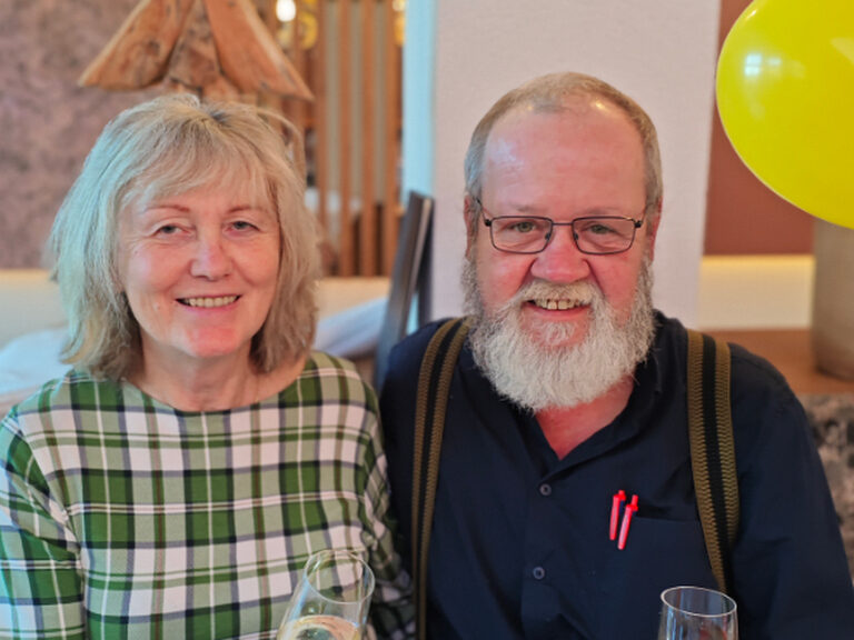 Paar stoßt anlässlich des Geburtstags der Frau mit einem Glas Sekt an, im Hintergrund sieht man einen gelben Luftballon
