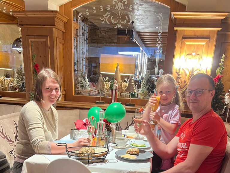 Familie sitzt am dekorierten Frühstückstisch; Eltern stoßen mit einem Glas Sekt und das Mädchen mit einem Glas Saft auf einen Geburststag an