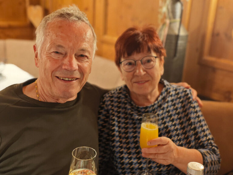 Paar sitzt am Geburtstagstisch, der Mann hat seinen Arm um die Frau gelegt und sie stoßen mit einem Glas Sekt bzw. einem Glas Orangensaft auf den Geburtstag an