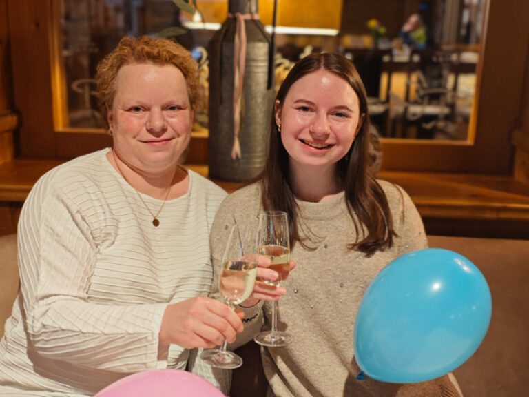 Zwei Frauen stoßen auf einen Geburtstag mit je einem Glas Sekt an und halten bunte Luftballone in der Hand