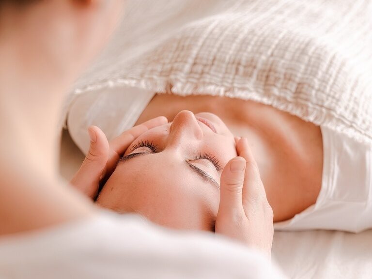Frau liegt in weißer Kleidung auf einer Liege und wird im Gesicht massiert