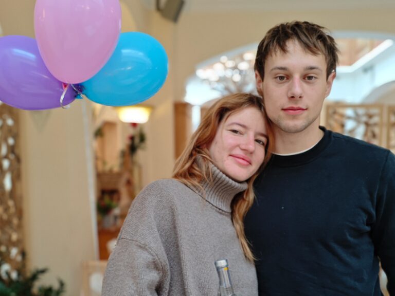 Paar posiert mit Geschenk anlässlich Geburtstag der Frau, im Hintergrund sind bunte Luftballons zu sehen