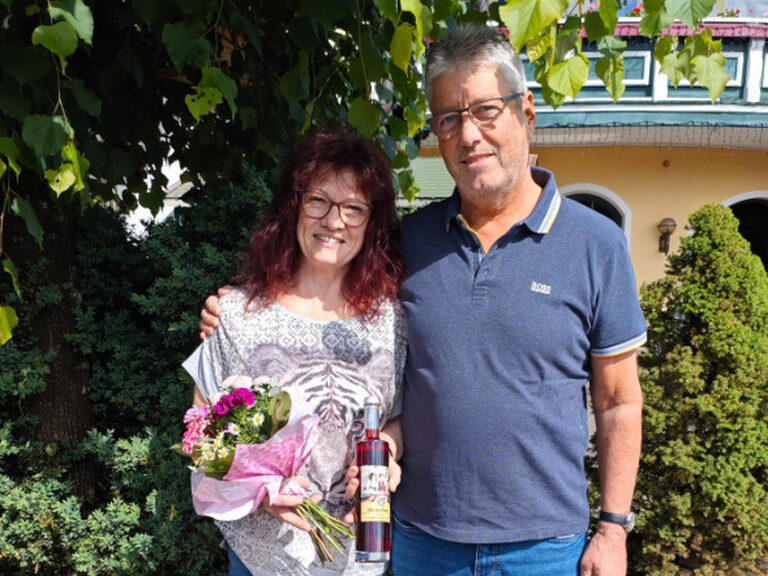 Paar steht im Romantikgarten und posiert lächelnd mit Blumenstrauß und Geschenk anlässlich des Geburtstags der Frau