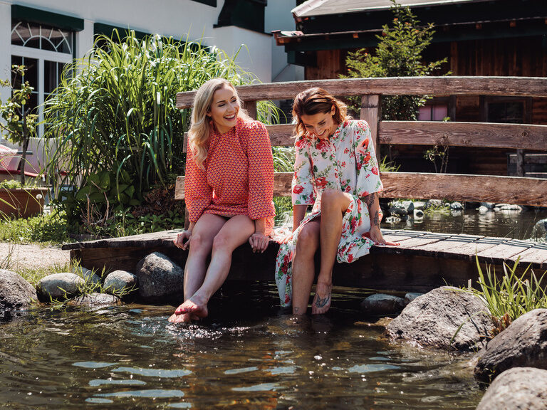 zwei Frauen sitzen in Sommerkleidern auf der kleinen Holzbrücke im Romantikgarten und lassen ihre Füße in den Teich baumeln