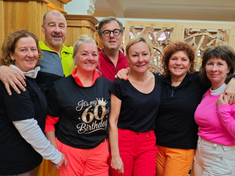 Eine Gruppe bestehend aus 7 Personen ist in farbenfrohen Skioutfits gekleidet und feiert einen 60. Geburstag