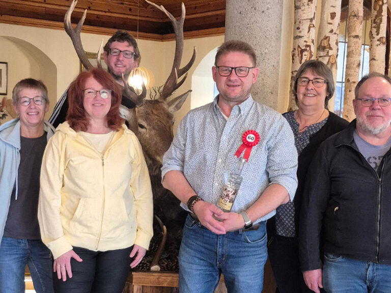 Drei Männer und drei Frauen posieren vor einem ausgestopften Hirsch mit einer Flasche Schnaps vom Hotel
