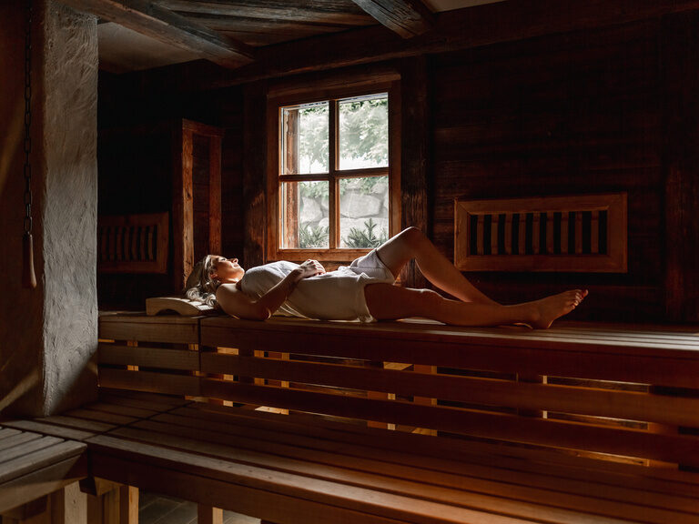 Frau liegt in einem Saunahandtuch gewickelt auf der Holzbank der Sauna und genießt einen Saunadurchgang