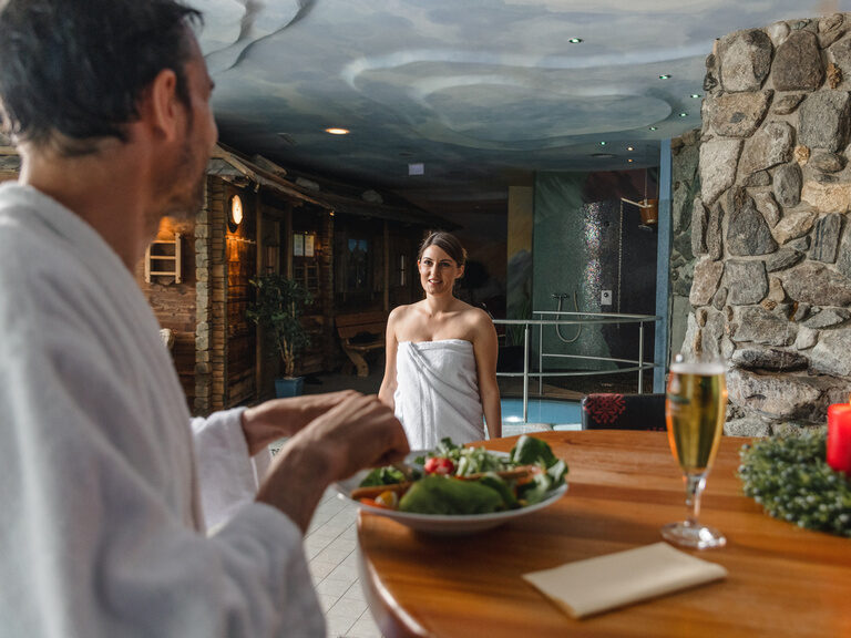 Paar im Barbereich der Saunawelt, der Mann isst an der Bar einen Salat und die Frau geht auf ihn zu
