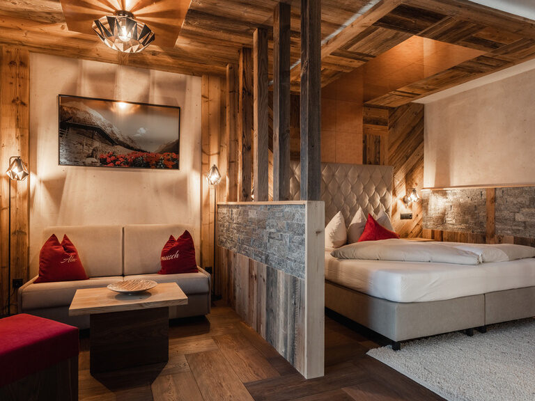 Suite Almrausch mit Boxspringbett, Holzvertäfelung, Holzboden mit grauem Hochflor-Teppich und gemütlicher Sitzecke hinter einem Raumteiler