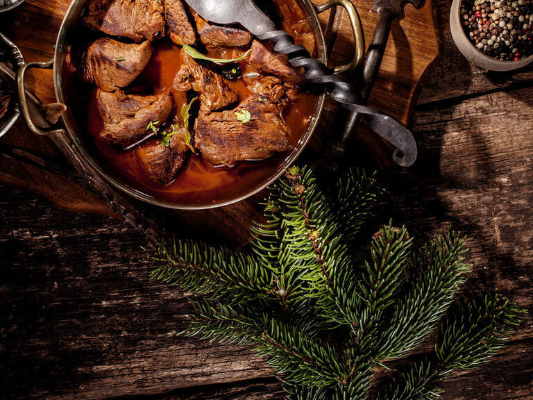 Gusseiserne Pfanne gefüllt mit einem winterlichen Schmorgericht steht auf dem Tisch und rundherum liegen Latschen als Deko