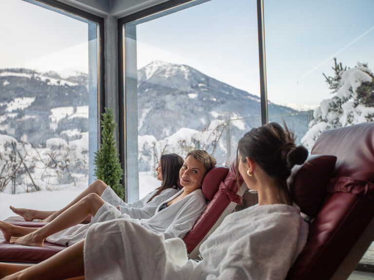 drei Frauen im Bademantel entspannen auf den Liegen im Ruheraum und genießen den Ausblick auf die verschneite Bergwelt