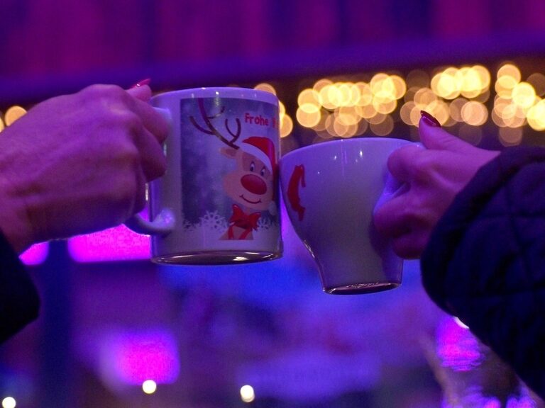 zwei weihnachtliche Tassen, mit denen angestoßen wird, und im Hintergrund ist Weihnachtsbeleuchtung zu sehen